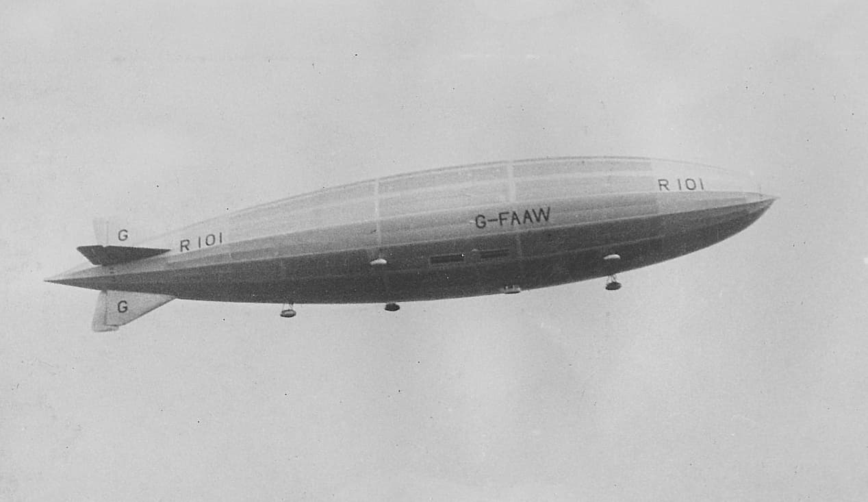 A black‑and‑white photograph of the British airship R101 in flight against a light, cloudy sky. The long, streamlined hull is shown side‑on, displaying the markings “R101,” “G‑FAAW,” and the tail identifier “G.” The airship’s fins and gondolas are visible beneath the envelope, and the craft appears to be cruising smoothly at altitude with no ground features in view.