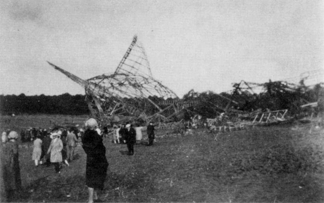 Black‑and‑white photograph showing the wreckage of a large airship collapsed across an open field. The metal framework lies twisted and broken, with sections of the structure bent at sharp angles and scattered across the ground. Groups of onlookers stand nearby, some in coats and hats, observing the devastation from various distances. Trees line the horizon under an overcast sky, giving the scene a somber, quiet atmosphere.