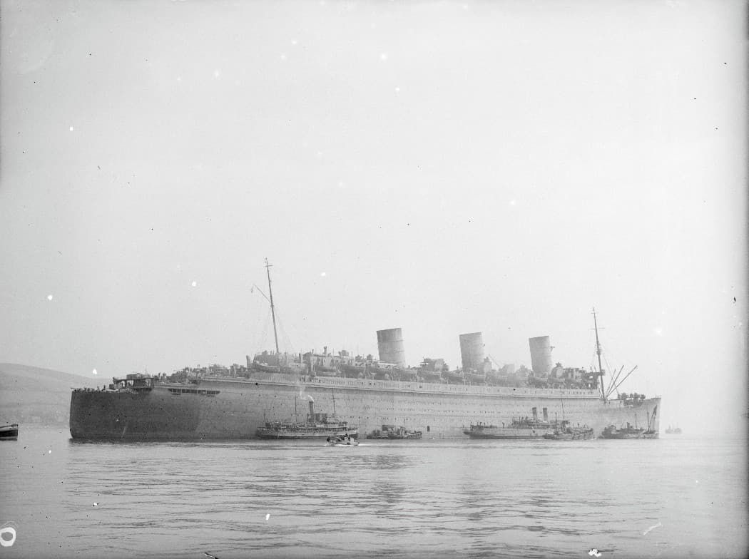 Black‑and‑white photograph of a large ocean liner converted for use as a troopship, anchored in a calm bay. The ship has a long hull and three tall funnels, with lifeboats and fittings mounted along the upper decks. Several small craft and tugboats cluster around the vessel near the waterline. The distant shoreline is faintly visible through light haze under an overcast sky.