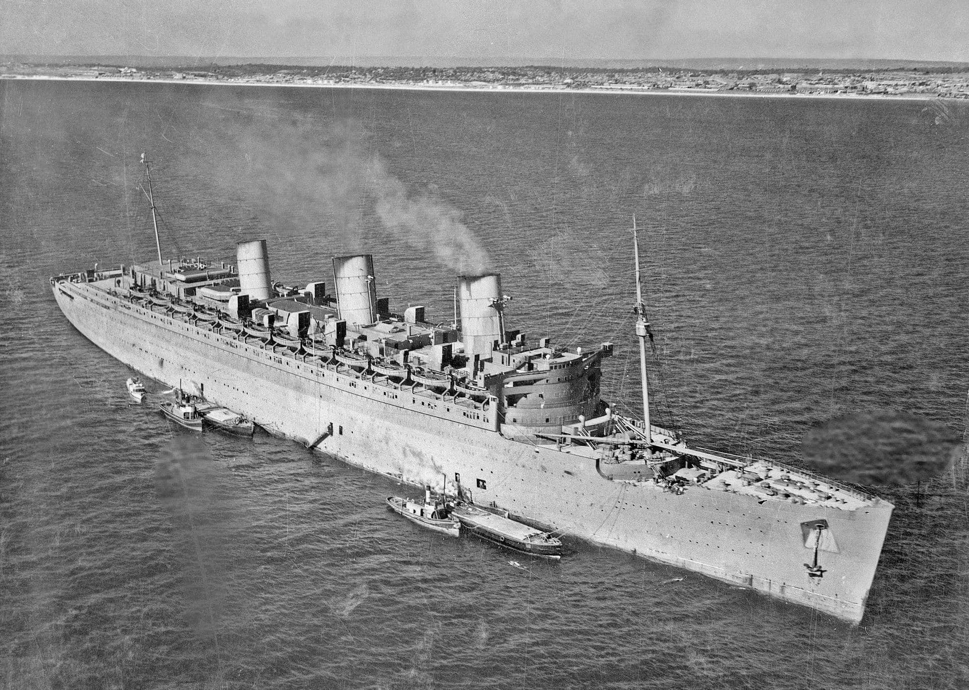 Black‑and‑white aerial photograph of a large troopship with three funnels anchored offshore in calm water. Tugboats and smaller service craft are positioned along the ship’s starboard side. Smoke rises lightly from the funnels, and the vessel’s decks appear crowded with equipment and lifeboats. In the distance, a low coastline stretches across the horizon beneath a clear sky