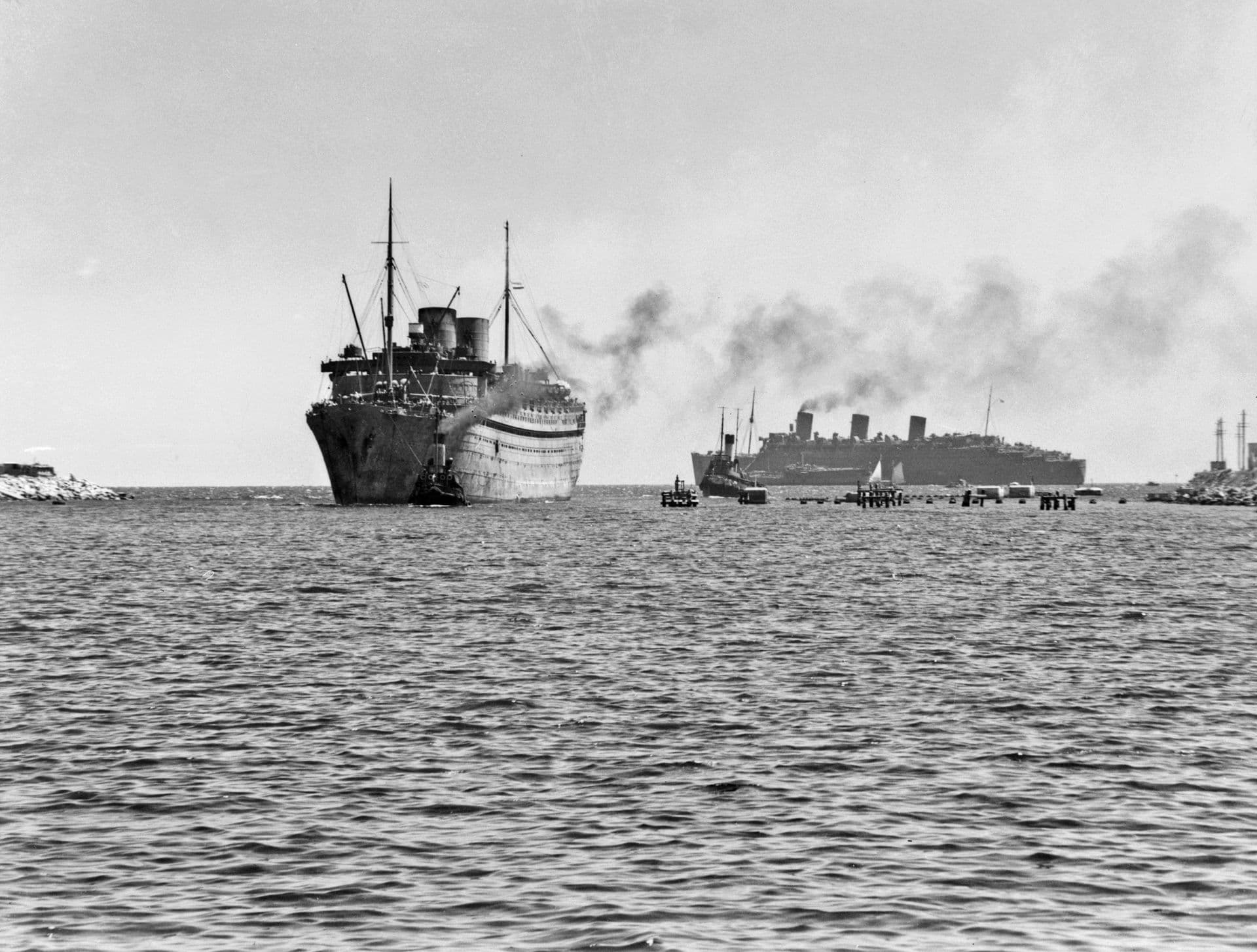 Black‑and‑white photograph showing two large passenger liners operating as troopships during wartime. In the foreground, a ship moves slowly toward the camera with a tug positioned at its bow. Another large liner sails behind it, trailing a plume of dark smoke across the sky. Smaller boats and harbour structures are visible near the waterline, and the scene is set on a calm sea under a lightly hazy sky.