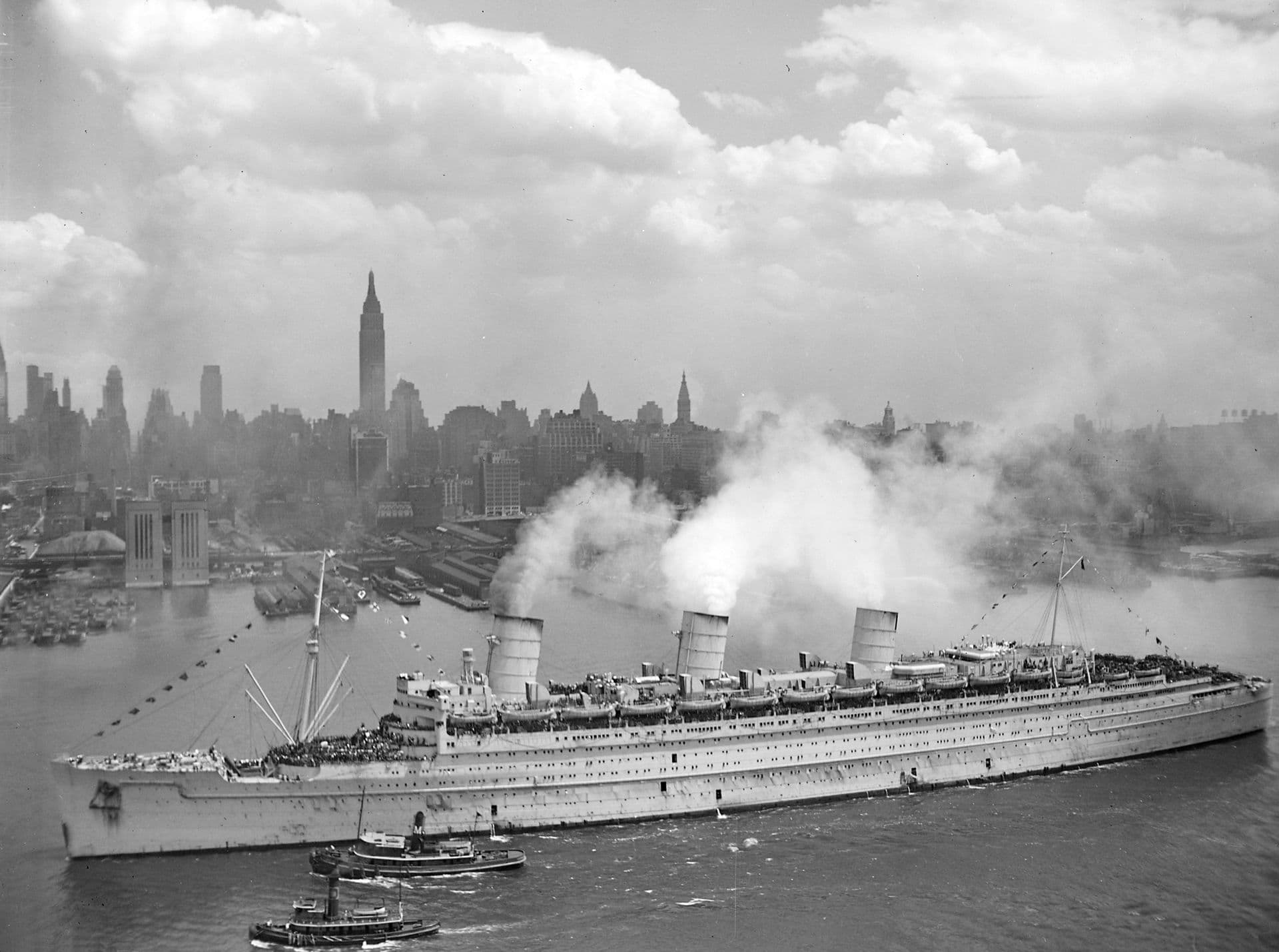 A black‑and‑white aerial photograph of a large ocean liner moving through a wide harbour channel. The ship has three tall funnels releasing plumes of white steam, and its long decks are lined with equipment and lifeboats. Several tugboats travel alongside and near the bow. In the background, the skyline of a major city rises beneath a partly cloudy sky, with tall buildings and piers visible along the waterfront.