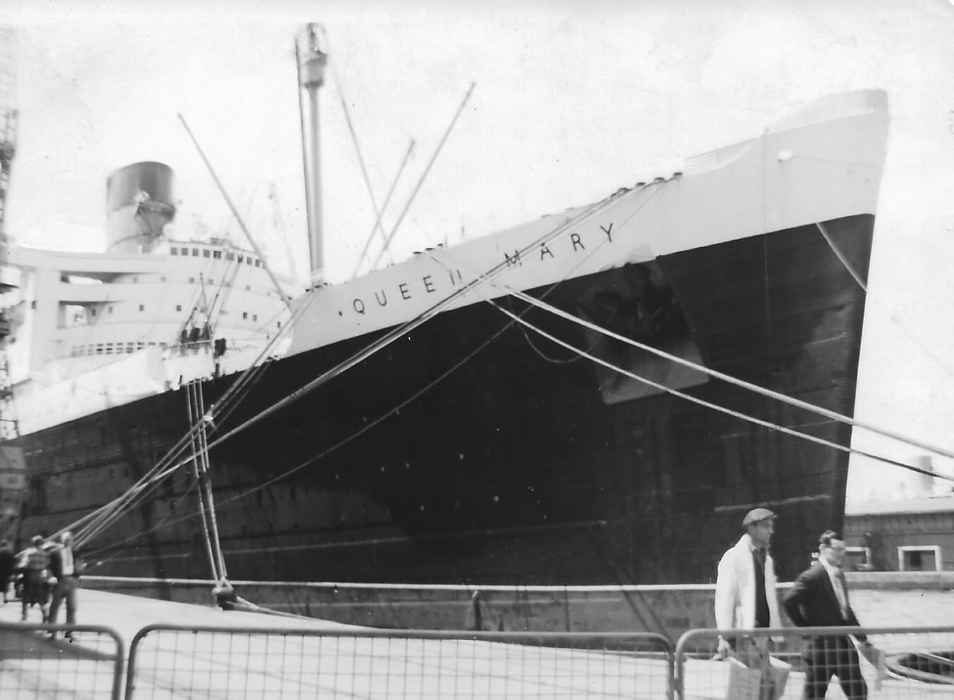 A black‑and‑white photograph showing the bow of the ocean liner Queen Mary moored at a wharf. Thick mooring lines stretch from the ship down to bollards on the dock. The towering hull dominates the frame, with the ship’s name visible near the bow. A few people walk along the waterfront in the foreground, while parts of the ship’s superstructure and a single funnel rise above the deck under an overcast sky