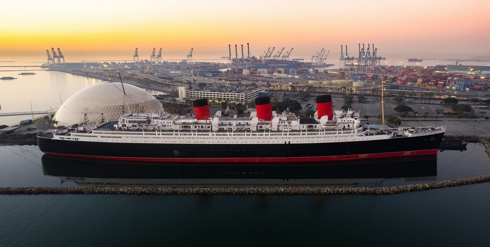 A wide, high‑angle colour photograph of the historic ocean liner Queen Mary docked in calm harbour waters at sunset. The ship’s three red‑and‑black funnels stand out along its long white superstructure and dark hull. A curved breakwater runs parallel to the vessel, and behind it stretches a busy port filled with cranes, shipping containers, and industrial buildings. The sky glows with soft orange and pink tones as daylight fades over the harbour.
