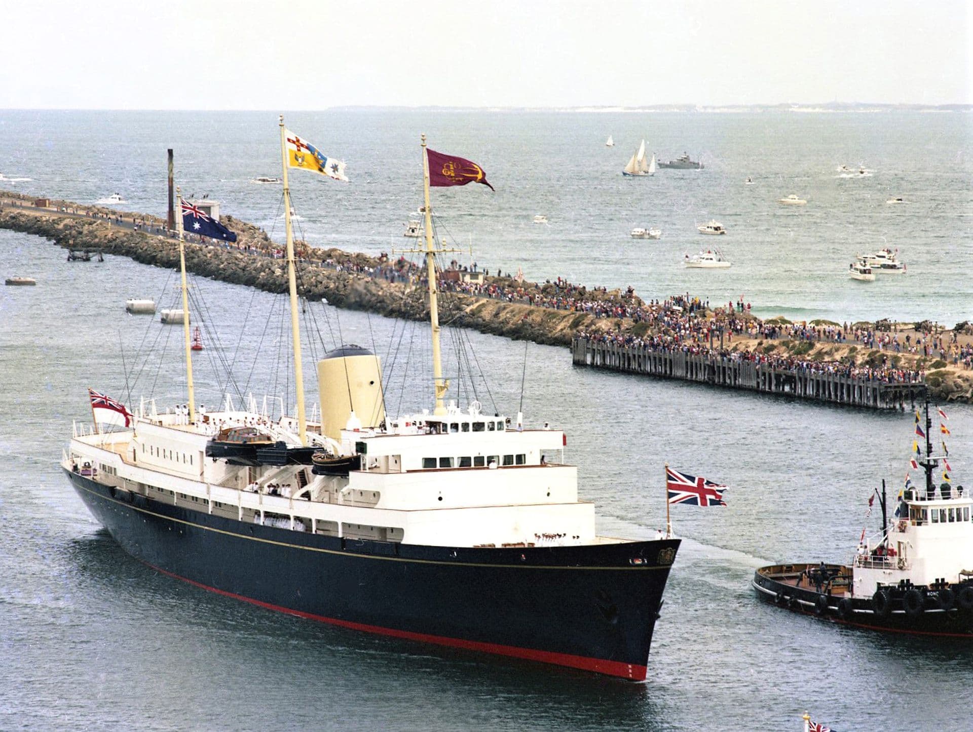 Colour photograph of a large royal yacht entering a harbour channel, accompanied by a tugboat. The vessel has a dark blue hull with a white superstructure and a single cream‑coloured funnel. Several ceremonial flags fly from tall masts along the deck, and a Union flag is displayed at the stern. Crowds line the breakwater in the background, watching the yacht’s arrival, while numerous small boats and yachts dot the surrounding water on a bright, calm day.