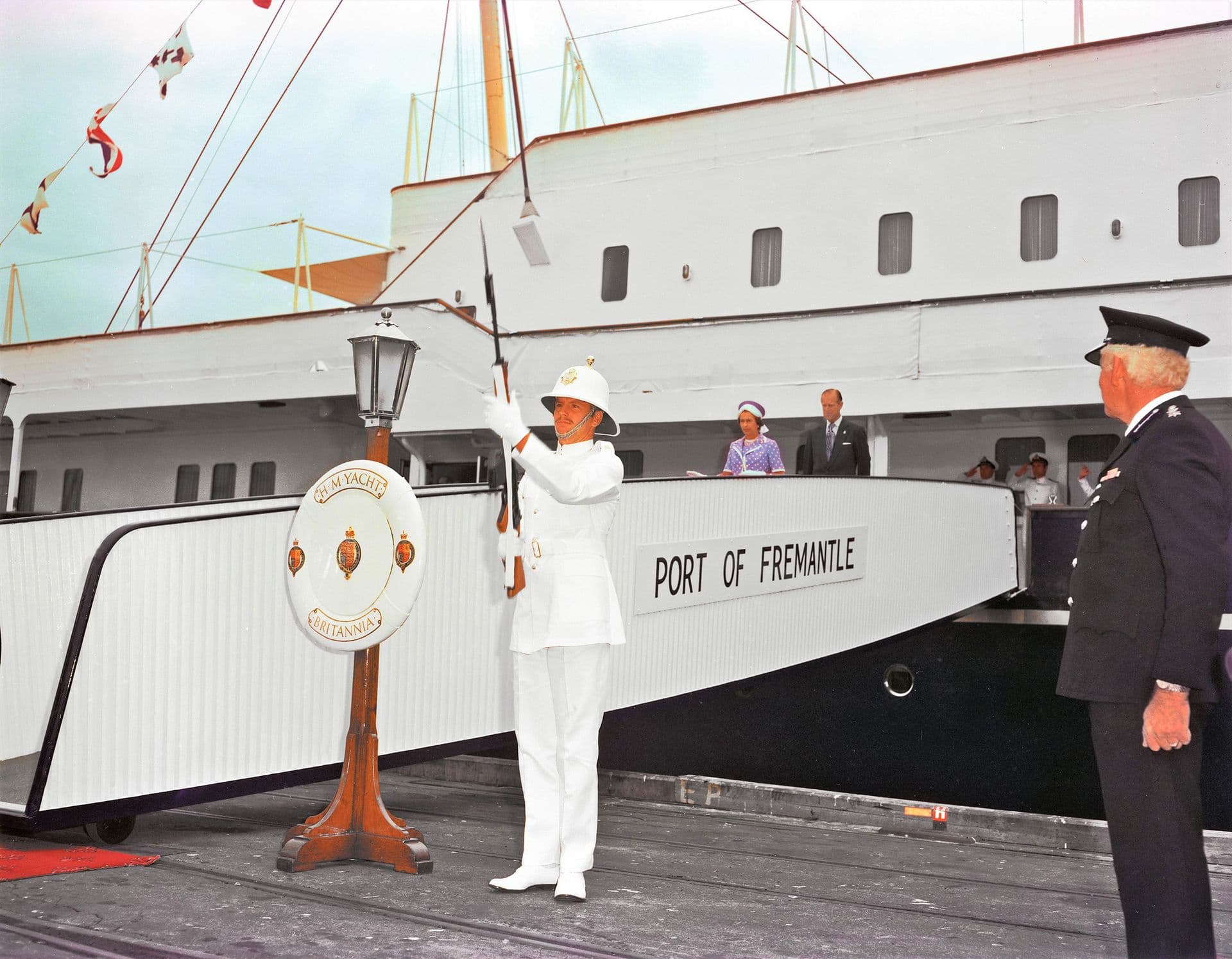 Colour photograph showing a ceremonial moment beside a royal yacht moored at the Port of Fremantle. In the foreground, a uniformed naval guard stands on the dock raising a rifle in a formal salute, positioned beside a decorative stand bearing an emblem. To the right, another uniformed official watches the ceremony. On the yacht’s gangway, two dignitaries stand side by side, observing the proceedings. The ship’s white hull and polished fittings form the backdrop, with signal flags strung above the deck.