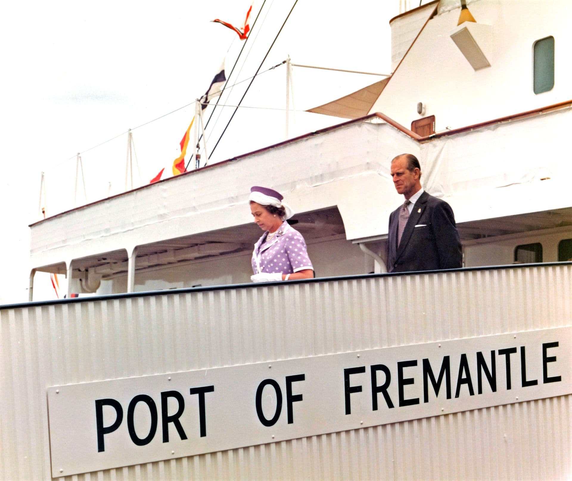 Colour photograph showing two well‑dressed dignitaries standing on the gangway of a large ship as it is docked at the Port of Fremantle. One is wearing a light-coloured dress and hat, while the other is dressed in a dark suit. Colourful signal flags hang along the ship’s railings above them. The metal gangway in the foreground has a large sign reading “PORT OF FREMANTLE,” and the ship’s white superstructure forms the backdrop.
