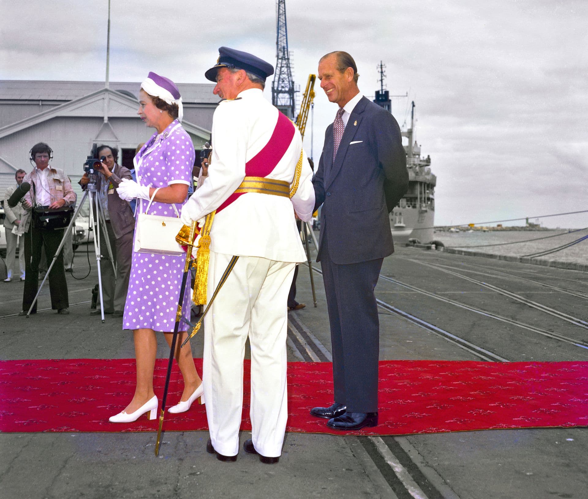 Colour photograph of three individuals standing on a red ceremonial carpet laid out on a dockside. One person wears a purple patterned dress with matching hat, gloves, and handbag. Another person beside them is dressed in a dark formal suit with hands clasped behind their back. A third person, wearing a white dress uniform with a blue cap and ceremonial sash, stands facing them while holding an ornate staff. Photographers and onlookers stand in the background near buildings and harbour structures, and a lar