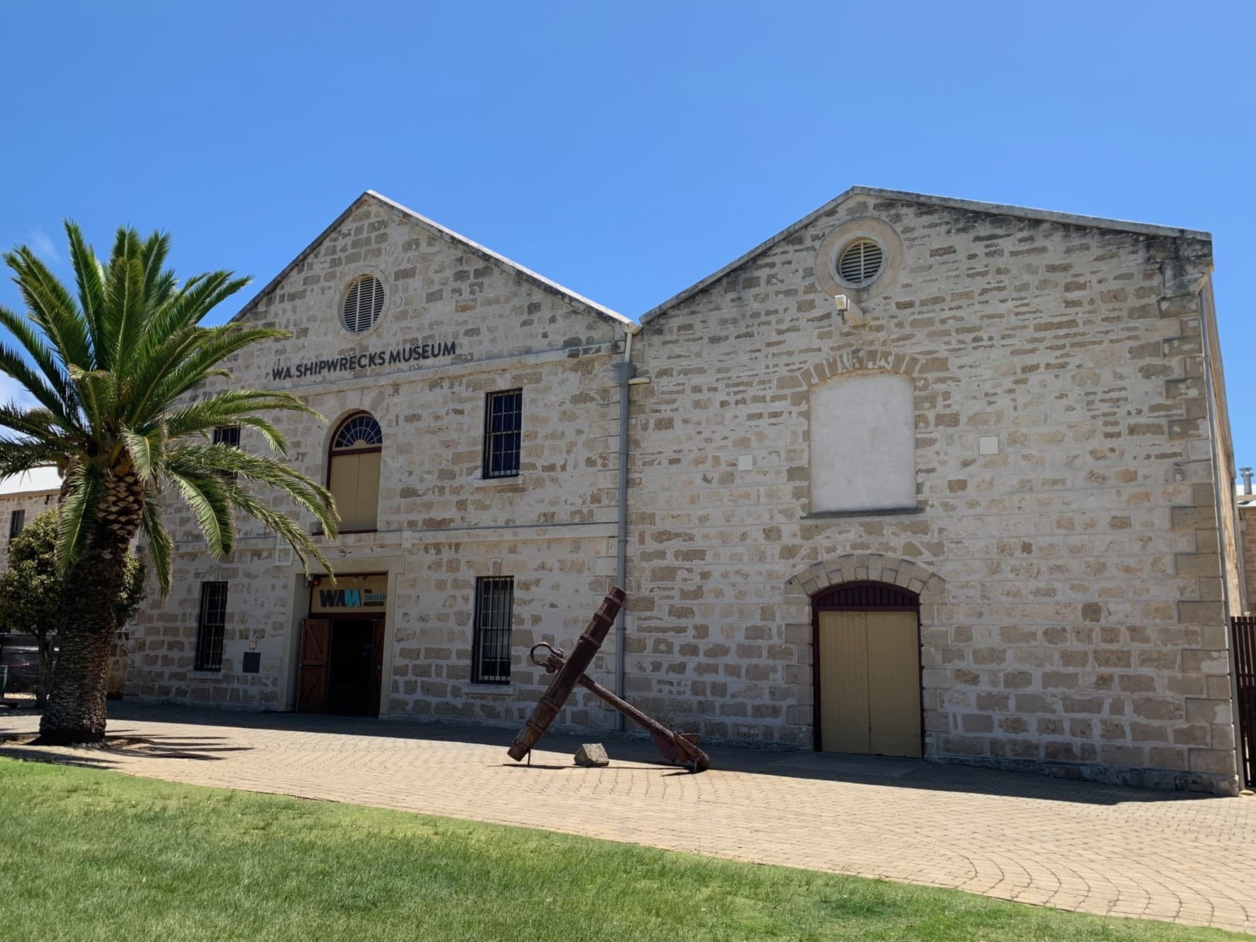 A two-story limestone building, with a paved entry way. A large anchor sits on the pavement. In front of the building is a large palm tree, and a green grass lawn.