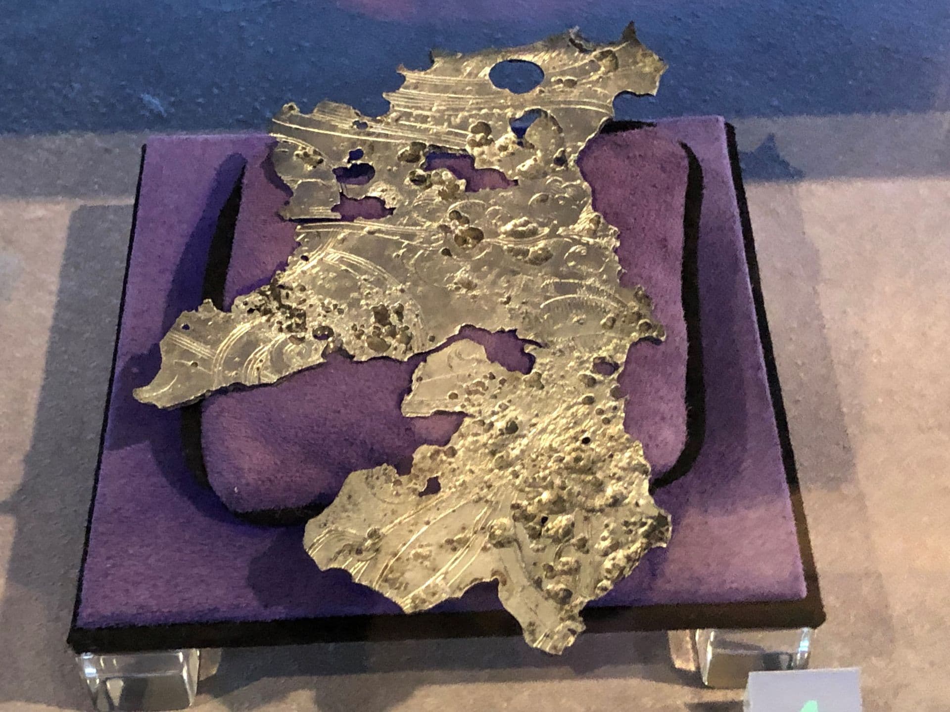 Jagged piece of silver metal with holes in it placed on a purple cushion in a display case.