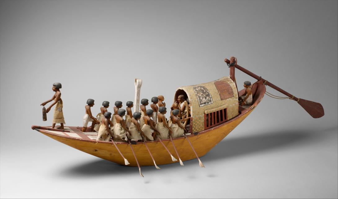 Small model of a boat with figurines of 14 people rowing and one person standing a the front of the boat.