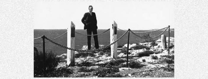 Black and white photo of man in a suit standing on a cliff overlooking the ocean with two posts surrounded by chain bollards.